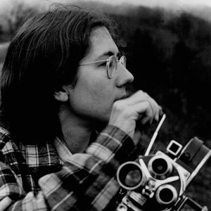 The image is a black and white photograph depicting a young person with medium-length hair, wearing round glasses and a plaid shirt. They are in a contemplative pose, resting their cheek on one hand, while holding a Bolex camera with the other hand. The camera is pointed towards the left of the frame and has two lenses visible. The person appears to be lost in thought, gazing into the distance. The background is out of focus, suggesting a natural outdoor setting. The image exudes a vintage feel, likely from the early 1970s, and captures a moment of introspection, possibly between film shoots.